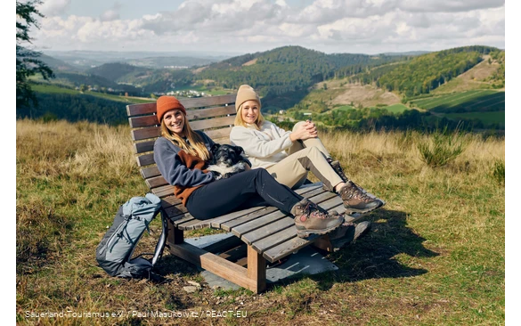 Zwei Frauen und ein Hund machen eine Pause auf einer Bank mit weiter Aussicht