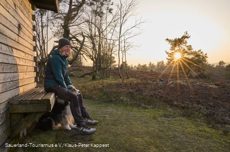 Auf einer Bank an einer Hütte auf dem Kahlen Pön sitzt Frau mit Hund im Winterlicht